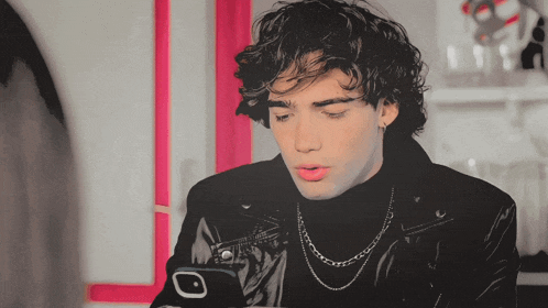 a young man wearing a black turtleneck and a silver chain looks at his phone
