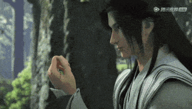 a man with long black hair is holding something in his hand in a forest .