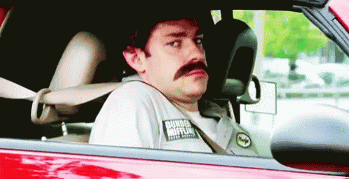 a man with a mustache is driving a red car and wearing a dunkin donuts shirt .