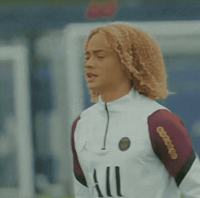 a woman with curly hair is wearing a white and maroon soccer jersey and standing on a field .