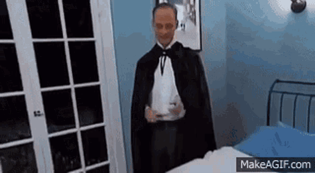 a man in a tuxedo and cape is standing in a bedroom next to a bed .