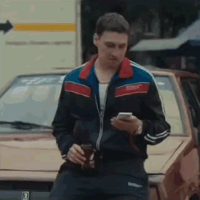 a man is sitting in front of a car looking at his phone while holding a cup of coffee .