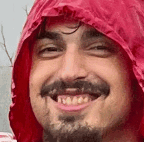 a man with a beard and mustache is wearing a red hooded jacket and smiling .