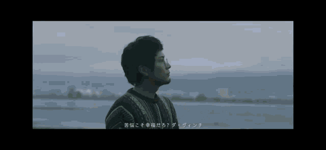 a man in a sweater stands in front of a body of water with a city in the background