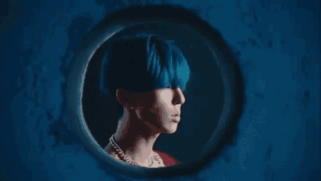 a man with blue hair and a pearl necklace is looking through a hole in a wall .