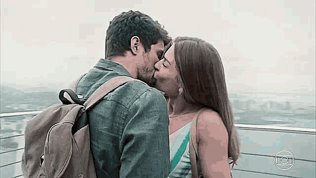 a man and a woman are kissing on a balcony . the man is wearing a backpack .
