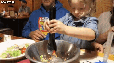 a little girl is mixing a salad in a bowl with a spoon while a man in a california shirt watches .