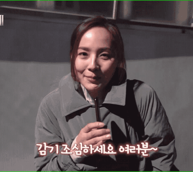 a woman in a grey coat is smiling and holding a cell phone with korean writing on the bottom right