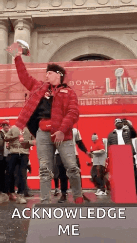 a man in a red jacket is standing on a stage holding a football and a trophy .