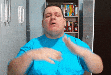 a man in a blue shirt is making a gesture with his hands in front of a bookshelf with bottles of hot sauce