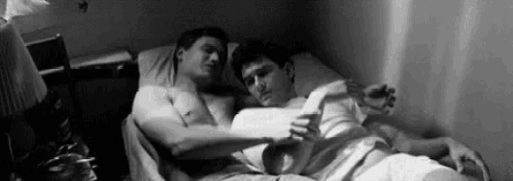 a black and white photo of two men laying on a bed . one of the men is reading a book .