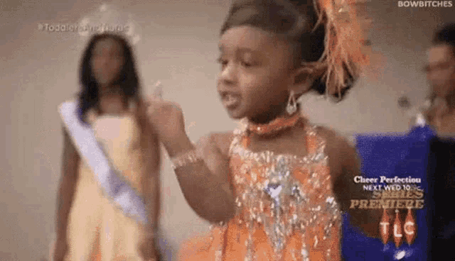 a little girl in an orange dress is giving the middle finger to another little girl .