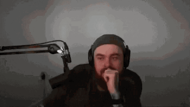 a man with a beard is sitting in front of a microphone wearing headphones and a hat .