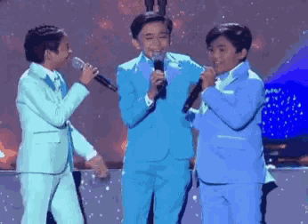 three young boys in blue suits singing into microphones on a stage