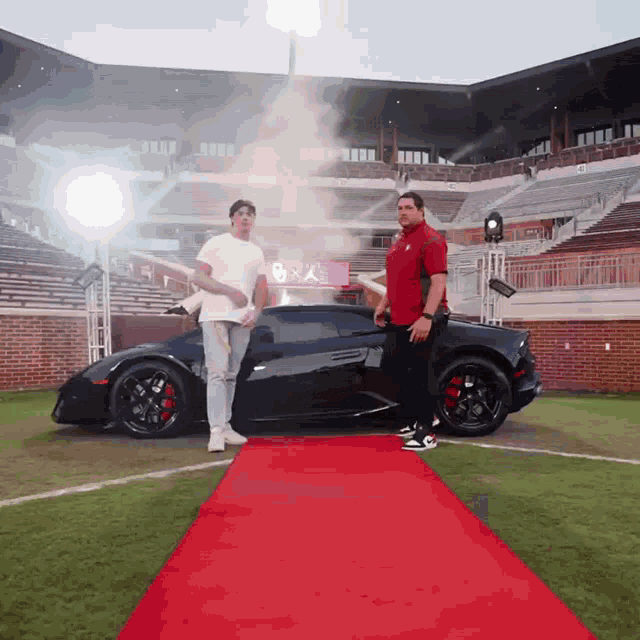 two men standing next to a black sports car on a red carpet in front of a stadium