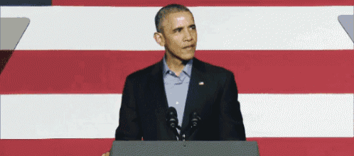 a man in a suit stands at a podium in front of an american flag