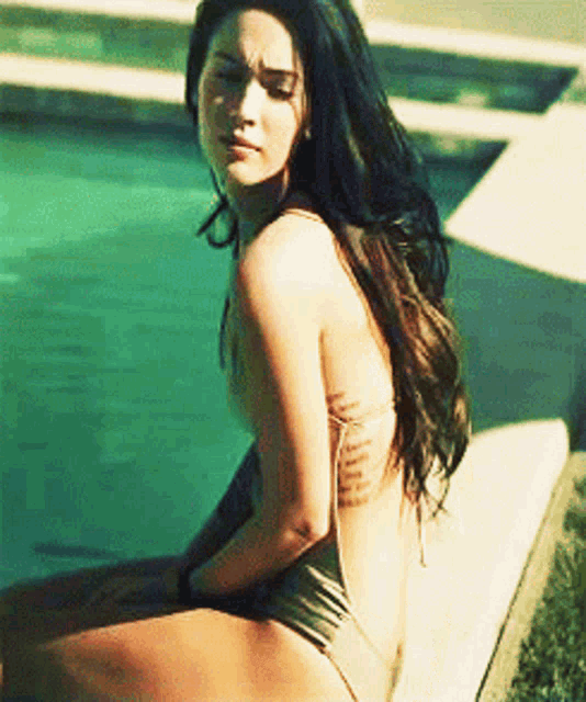 a woman in a bikini sits on a ledge near a pool