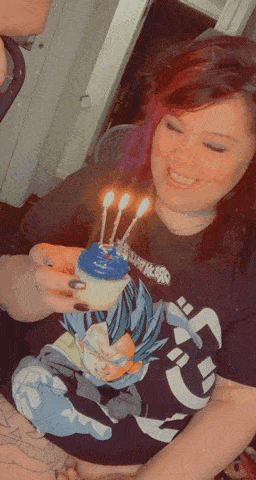 a woman in a dragon ball z shirt is holding a cupcake with three lit candles .