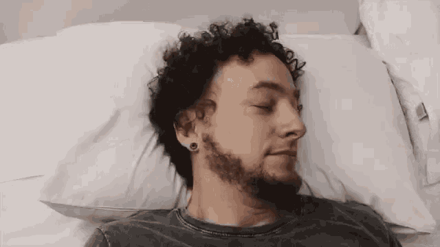 a man with curly hair and a beard is sleeping in a bed with his eyes closed .