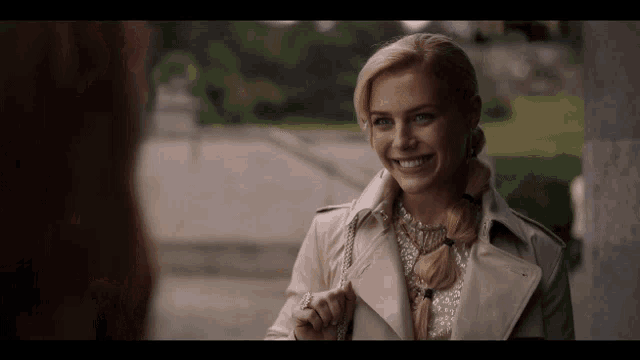 a woman wearing a trench coat and a braided ponytail is smiling
