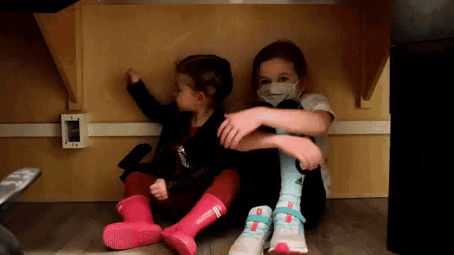 two little girls wearing face masks sit under a table