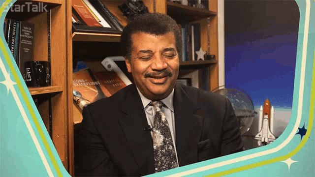 a man in a suit and tie is smiling in front of a bookshelf with a book titled signs and signs of the zodiac