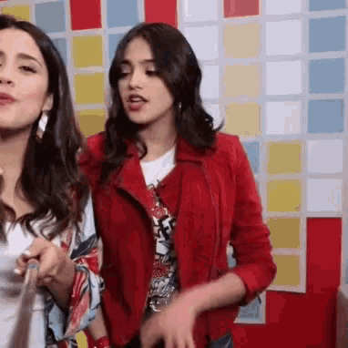 two women are standing next to each other in front of a colorful wall . one is wearing a red jacket .