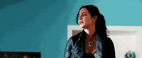 a woman wearing a leather jacket and a necklace is standing in a room with a blue wall .
