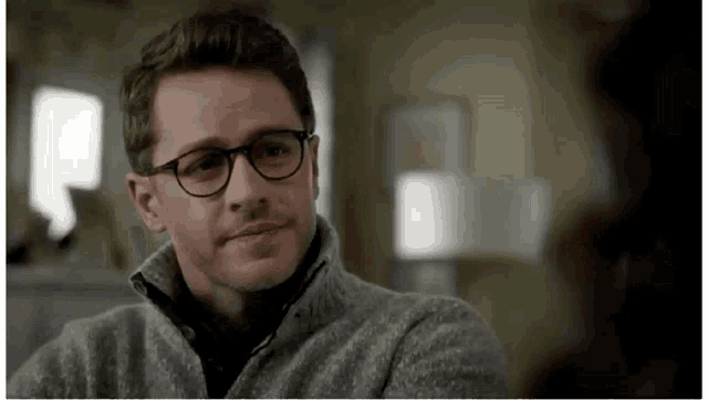 a man wearing glasses and a grey sweater is sitting in a room .