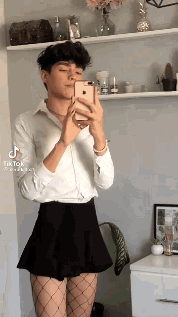 a young man is taking a selfie with his cell phone while wearing a skirt and fishnet stockings .