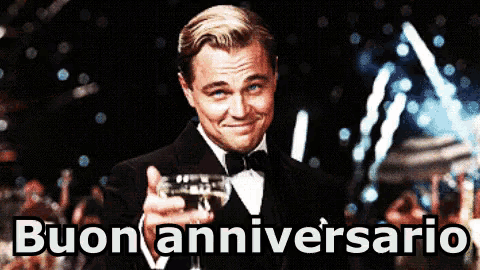 a man in a tuxedo is holding a glass of wine in front of a sign that says buon anniversary