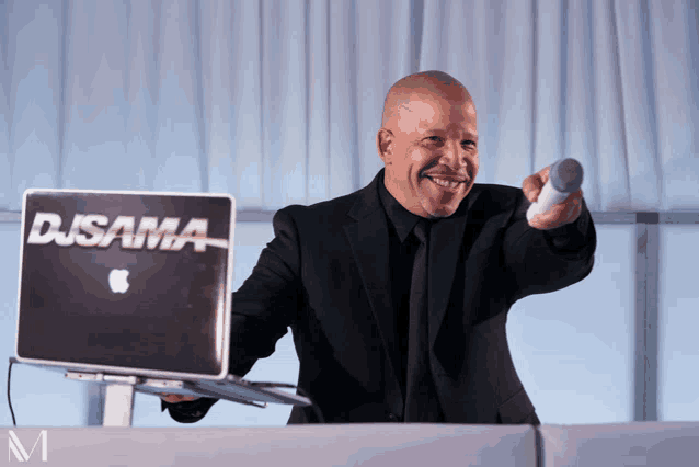 a man in a suit holds a microphone in front of a djsama laptop