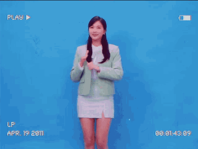 a woman in a green jacket and a white skirt is standing in front of a blue background .