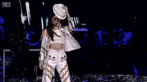 a woman wearing a white hat and a crop top stands on a stage with a microphone in front of her