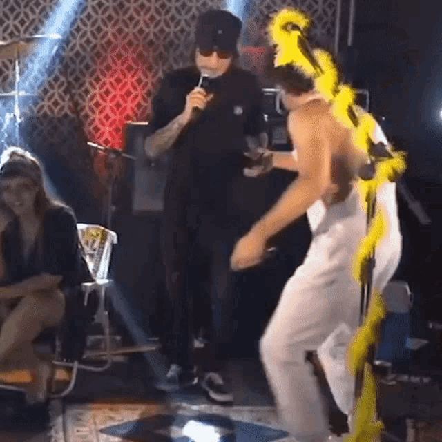 a man wearing a yellow feathered headband is dancing on a stage while another man sings into a microphone .