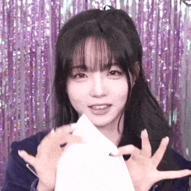 a young woman with long black hair and bangs is holding a piece of paper in front of her face .
