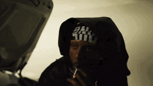 a man in a hooded jacket is smoking a cigarette next to a car with the hood up .