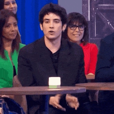 a man in a suit and glasses is sitting at a table in front of a crowd of people .