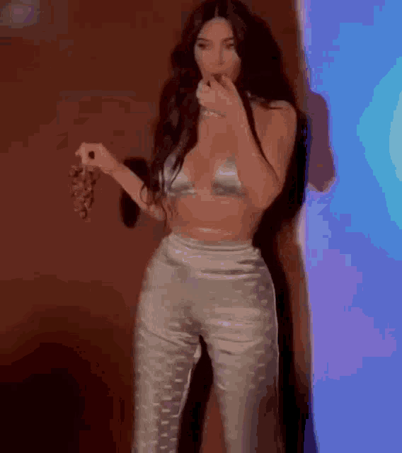a woman in a crop top and pants is standing next to a wall holding a bunch of grapes .