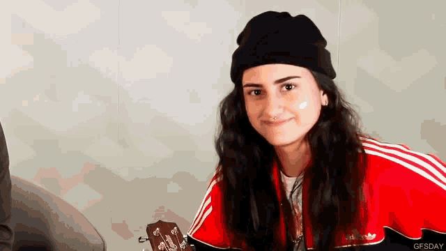 a woman wearing a black beanie and a red adidas jacket is holding a guitar