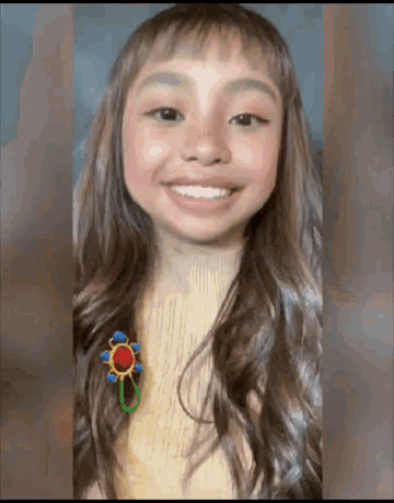 a young girl with long hair and bangs is smiling and wearing a brooch .