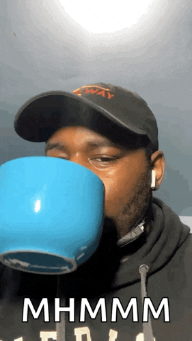 a man wearing a hat and headphones is drinking from a blue cup with the words mhmmmm written on it