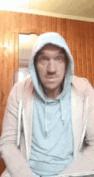 a man in a hoodie is sitting in front of a wooden wall and making a funny face .