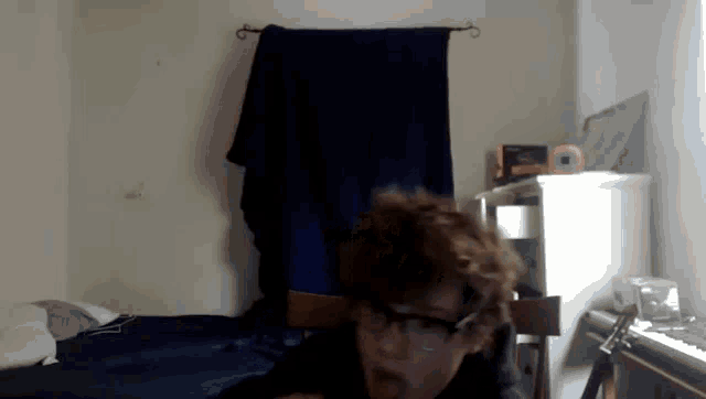 a person wearing glasses is sitting in a room with a piano and a blue blanket hanging on the wall .