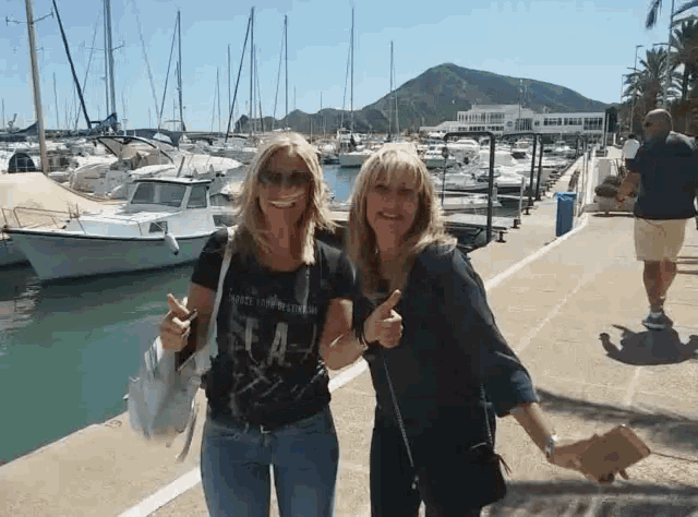 two women giving a thumbs up in front of a marina with boats