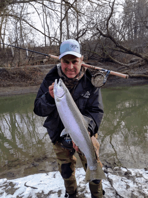 a man holding a rainbow trout in front of a river with a fishing rod