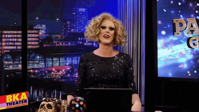 a drag queen stands in front of a screen that says bka theater