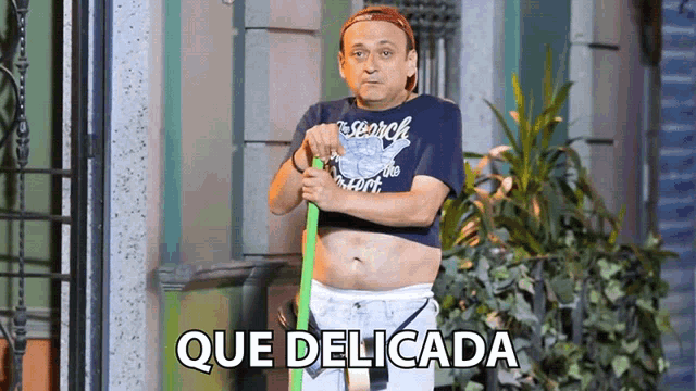a man wearing a search shirt is holding a green broom and says que delicada on the bottom