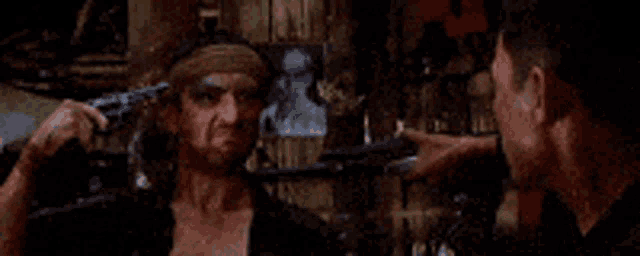 a man with a bandana on his head is pointing at another man with a gun .