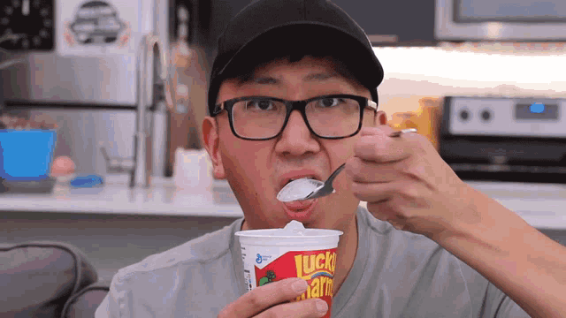 a man wearing glasses and a hat is eating a lucky charms cup with a spoon